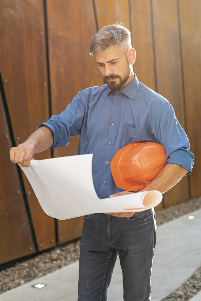 Mężczyzna z planem architektonicznym i pomarańczowym kaskiem pod pachą przegląda projekt budynku na tle nowoczesnej elewacji"